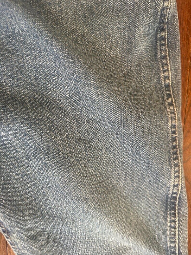 Puede incluir: Primer plano de tejido vaquero azul claro. El denim tiene un aspecto texturizado con detalles de tejido visibles. Una costura recorre verticalmente el lado derecho, con una puntada en contraste. El tono general es descolorido y desgastado.