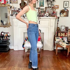 Puede incluir: Una persona con un top cors&eacute; verde claro con ribete floral, vaqueros azules y botas negras. La persona tambi&eacute;n lleva una gorra de b&eacute;isbol negra con texto dorado. El fondo incluye una chimenea y varios objetos decorativos.
