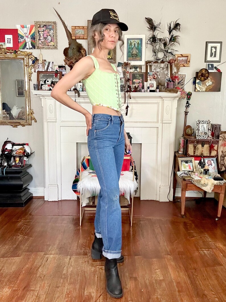Puede incluir: Una persona con un top cors&eacute; verde claro con ribete floral, vaqueros azules y botas negras. La persona tambi&eacute;n lleva una gorra de b&eacute;isbol negra con texto dorado. El fondo incluye una chimenea y varios objetos decorativos.