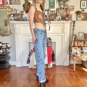 Puede incluir: Un top corto con estampado floral y cordones en la parte delantera, combinado con vaqueros de mezclilla de lavado claro y botas de plataforma negras. El top presenta un fondo marr&oacute;n con grandes dise&ntilde;os florales rojos y amarillos. Los vaqueros est&aacute;n doblados en los tobillos.