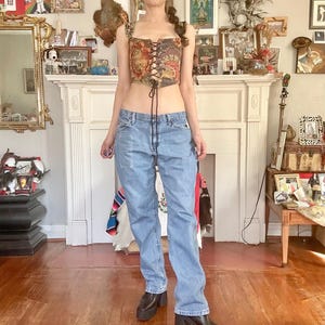 Puede incluir: Un top cors&eacute; de estilo vintage con dise&ntilde;o floral y tapiz, con cordones en la parte delantera. Combinado con vaqueros de mezclilla de lavado claro y botas de plataforma negras. El atuendo se presenta en una habitaci&oacute;n con chimenea y varios objetos decorativos.