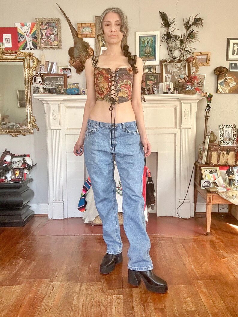 Puede incluir: Un top cors&eacute; de estilo vintage con dise&ntilde;o floral y tapiz, con cordones en la parte delantera. Combinado con vaqueros de mezclilla de lavado claro y botas de plataforma negras. El atuendo se presenta en una habitaci&oacute;n con chimenea y varios objetos decorativos.