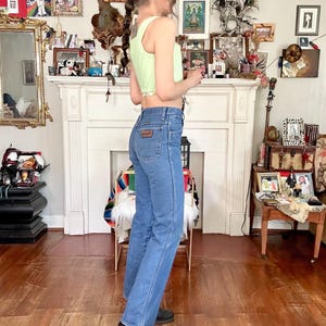 Puede incluir: Un top sin mangas verde claro y unos vaqueros azules con un parche Wrangler. Los vaqueros son de cintura alta y de pernera recta. La persona lleva un sombrero negro y botas negras. El fondo incluye una chimenea y varios objetos decorativos.