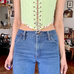 Puede incluir: Un cors&eacute; verde claro con cordones y ribete estampado, combinado con vaqueros azules. El cors&eacute; presenta una textura acanalada vertical y ojales plateados. Los vaqueros tienen un dise&ntilde;o cl&aacute;sico de cinco bolsillos.