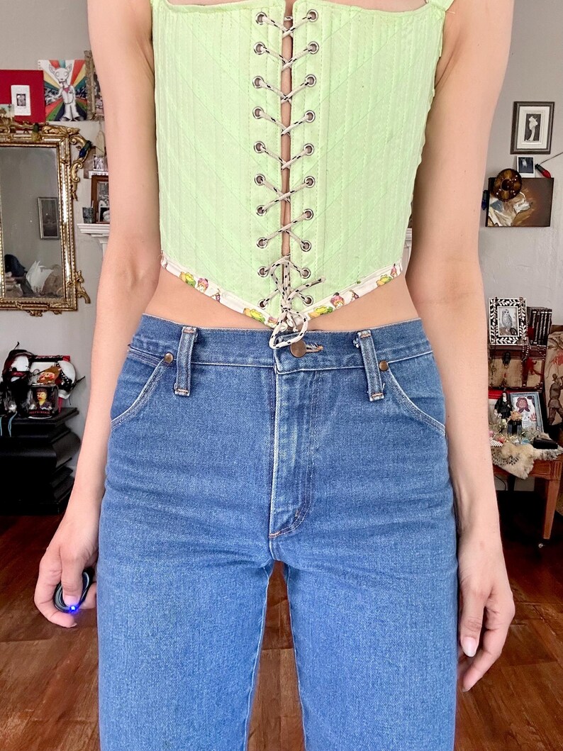 Puede incluir: Un cors&eacute; verde claro con cordones y ribete estampado, combinado con vaqueros azules. El cors&eacute; presenta una textura acanalada vertical y ojales plateados. Los vaqueros tienen un dise&ntilde;o cl&aacute;sico de cinco bolsillos.