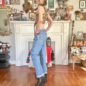 Puede incluir: Un top corto sin mangas con un estampado de cachemira en tonos marr&oacute;n, dorado y rojo, combinado con vaqueros de mezclilla azul claro y botas de plataforma negras. Los vaqueros est&aacute;n doblados en los tobillos.