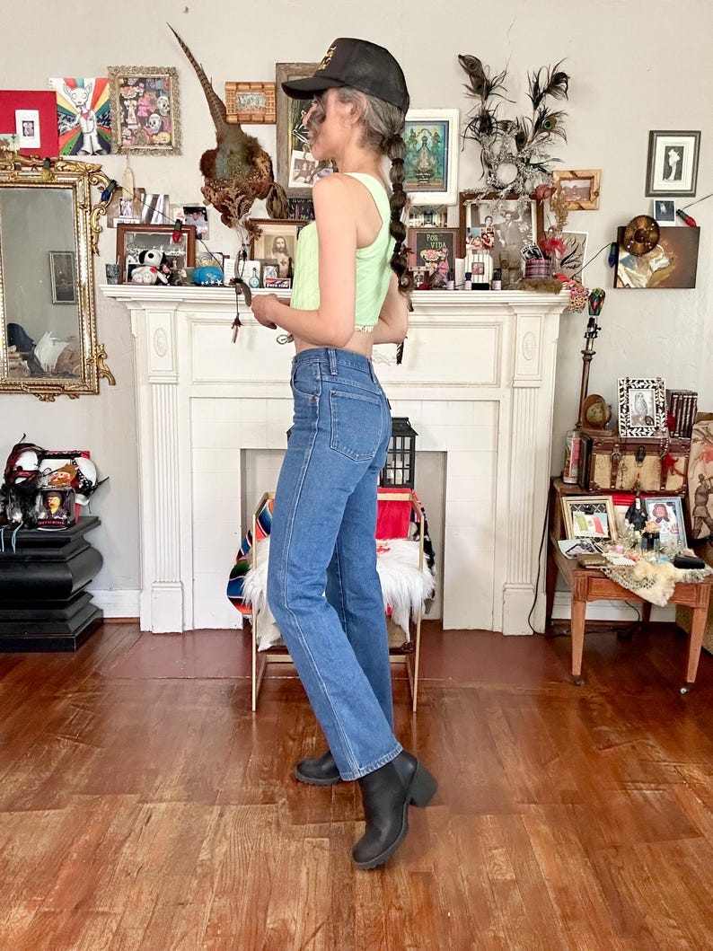Puede incluir: Un top corto acanalado verde claro combinado con jeans azules y botas negras. El atuendo se completa con una gorra de b&eacute;isbol negra. La modelo est&aacute; en una habitaci&oacute;n con chimenea y varios objetos decorativos.