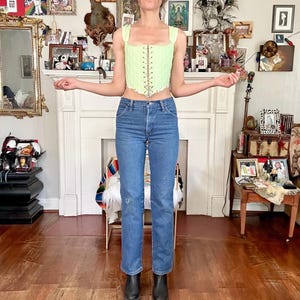 Puede incluir: Un top cors&eacute; verde claro con cuello cuadrado y cordones frontales se combina con vaqueros azules y botas negras. Se lleva una gorra de b&eacute;isbol negra con texto blanco. El fondo presenta una chimenea blanca y varias obras de arte enmarcadas.
