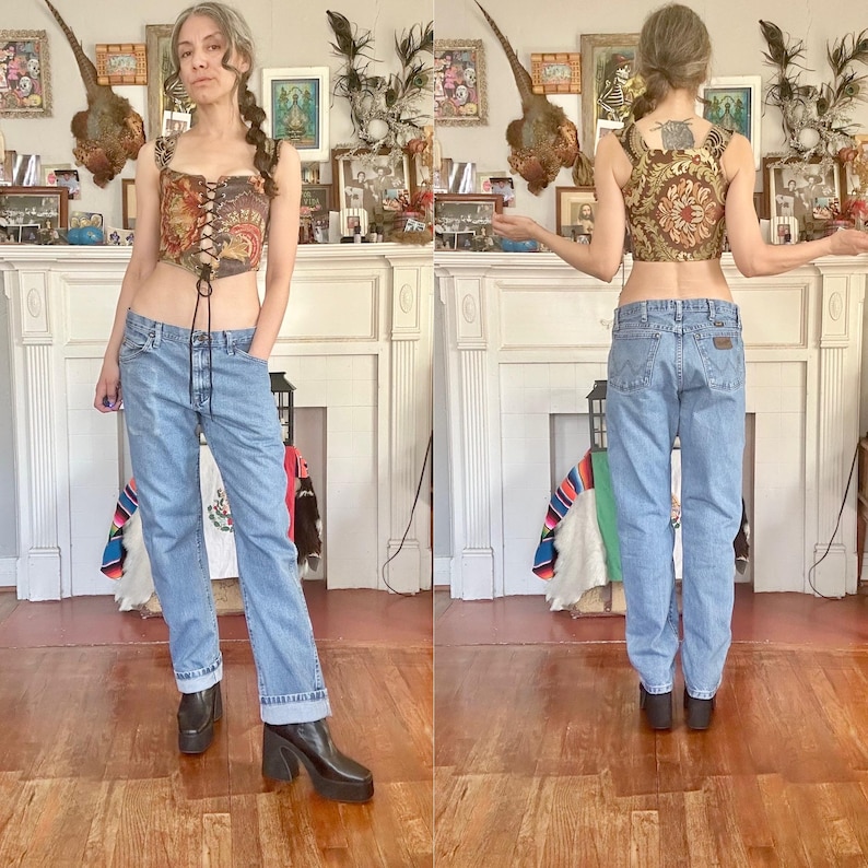 Puede incluir: Un conjunto de dos piezas con un top de cors&eacute; estampado y vaqueros de mezclilla de lavado claro. El cors&eacute; tiene un dise&ntilde;o floral marr&oacute;n y dorado con cordones negros. Los vaqueros est&aacute;n doblados en los tobillos. La persona lleva botas de plataforma negras.
