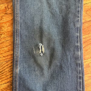 Puede incluir: Primer plano de una secci&oacute;n de vaqueros azules. Los vaqueros tienen un &aacute;rea desgastada con hilos deshilachados. La tela muestra signos de desgaste, con un aspecto descolorido. Las costuras son visibles a lo largo de las costurones.