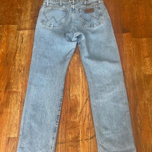 Puede incluir: Vaqueros de mezclilla azul claro con el logotipo de Wrangler en el bolsillo trasero. Los vaqueros tienen un dise&ntilde;o cl&aacute;sico de cinco bolsillos y un corte recto. Los vaqueros est&aacute;n colocados sobre una superficie de madera.