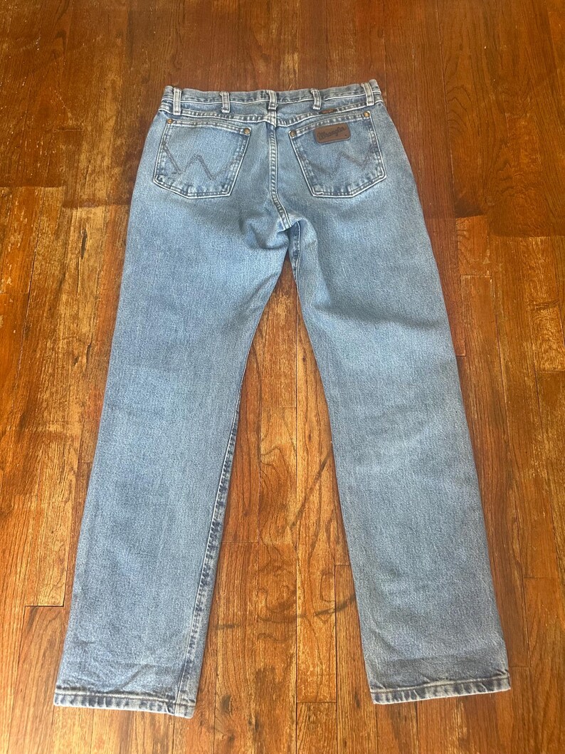 Puede incluir: Vaqueros de mezclilla azul claro con el logotipo de Wrangler en el bolsillo trasero. Los vaqueros tienen un dise&ntilde;o cl&aacute;sico de cinco bolsillos y un corte recto. Los vaqueros est&aacute;n colocados sobre una superficie de madera.