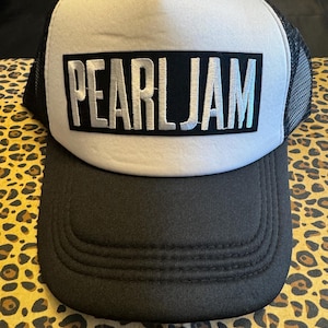Może przedstawiać: Czarno-biała czapka typu trucker z nazwą zespołu "PEARL JAM" w srebrnych literach na czarnym prostokącie. Czapka ma biały panel przedni, czarny daszek i czarny tył z siatki.