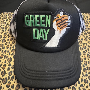 May include: Black trucker hat with a curved peak. The front displays a patch with the words "GREEN DAY" in green, and a cartoon goose holding a sandwich. The hat has a black front and a white and black plaid mesh back.