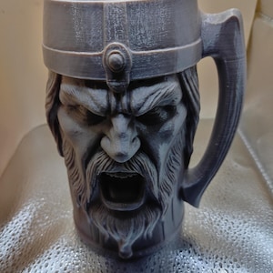 Può includere: Un boccale da birra grigio, stampato in 3D, con manico, raffigurante un volto vichingo arrabbiato e dettagliato. Il boccale ha un bordo largo e un manico grande. Il vichingo ha una lunga barba e indossa un elmo.