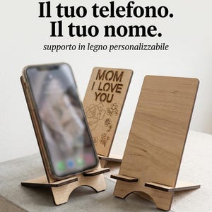 Può includere: Tre supporti per telefono in legno chiaro. Un supporto sostiene uno smartphone. Un altro supporto presenta il testo inciso "MOM I LOVE YOU" con un motivo floreale. La parte superiore dell'immagine mostra il testo "Il tuo telefono. Il tuo nome. supporto in legno personalizzabile" e la parte inferiore "DnyshpCreazioni".