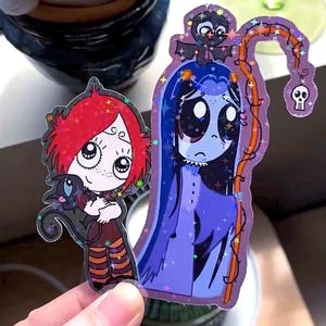 May include: Two holographic stickers featuring cartoon characters. One shows a girl with red hair holding a black cat. The other depicts a blue-skinned figure with long hair and a bat on its head, holding a vine with a skull.