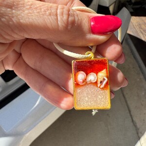 Puede incluir: Colgante rectangular con marco dorado, con un diseño de resina roja y beige. La resina encierra tres conchas marinas. El colgante cuelga de un cordón color crema.