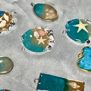 Puede incluir: Una colección de colgantes de resina hechos a mano con diseños temáticos del océano. Los colgantes presentan conchas marinas, estrellas de mar y resina azul y turquesa incrustadas, engastadas en marcos de metal plateado y dorado. Varias formas están presentes.