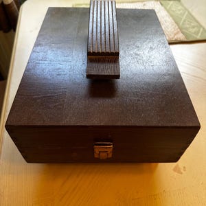 Puede incluir: Una caja de madera marrón oscuro para lustrar zapatos con reposapiés incorporado. La caja tiene forma rectangular con tapa con bisagras y cierre metálico. El reposapiés está hecho de listones de madera verticales.