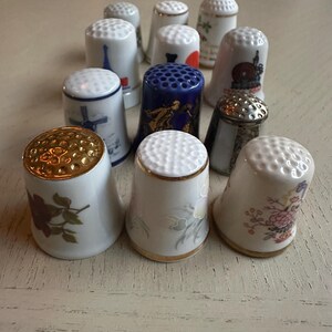 Könnte beinhalten: Eine Sammlung dekorativer Fingerhüte aus Keramik. Die Fingerhüte weisen verschiedene Designs auf, darunter Blumenmuster, Wahrzeichen und goldfarbene Akzente. Die Fingerhüte sind weiß, blau, gold und silberfarben. Sie sind auf einer hellen Oberfläche angeordnet.