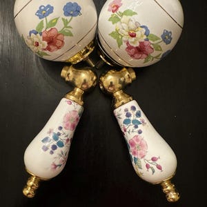 Könnte beinhalten: Zwei Sets weißer Keramik-Schrankknöpfe mit Blumendesigns und goldfarbener Hardware. Die Knöpfe weisen eine Mischung aus roten, blauen und rosa Blumen auf. Die Griffe haben eine tropfenförmige Form mit rosa und blauen Blumenmustern.