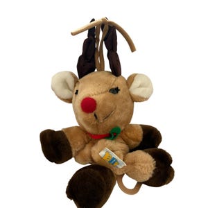 May include: A plush reindeer toy with light brown fur, dark brown hooves, and dark brown antlers. It features a bright red nose, white inner ears, and a red and green collar. A small white tag with a yellow bear icon is attached to the chest area.
