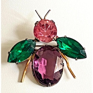 May include: A brooch shaped like an insect, featuring a round pink faceted stone for the head, an oval purple faceted stone for the body, and two marquise-cut green faceted stones for the wings, all set in a gold-toned metal frame with antennae.