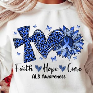 May include: White sweatshirt featuring a blue leopard print cross, heart, and sunflower design with a blue ribbon. The words "Faith Hope Cure" and "ALS Awareness" are printed below the graphic. Small blue butterflies are scattered around the design.