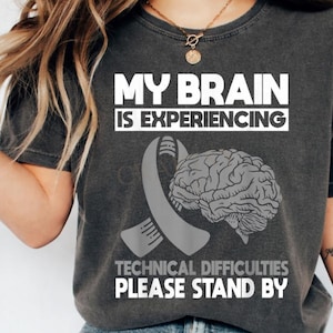 May include: Dark gray t-shirt with the text "MY BRAIN IS EXPERIENCING TECHNICAL DIFFICULTIES PLEASE STAND BY". The shirt features a gray ribbon and a detailed brain graphic.