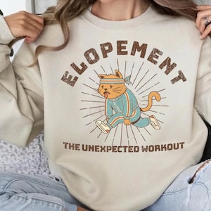 May include: Cream-coloured sweatshirt featuring the word "ELOPEMENT" above a cartoon cat in a headband and tracksuit. The cat has its tongue out. The text "THE UNEXPECTED WORKOUT" is below the cat.