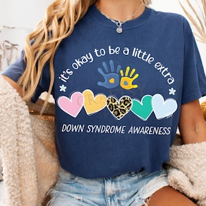Op de afbeelding: Marineblauw t-shirt met de tekst "it's okay to be a little extra" en "Down Syndrome Awareness". Het ontwerp bevat kleurrijke harten en handafdrukken.