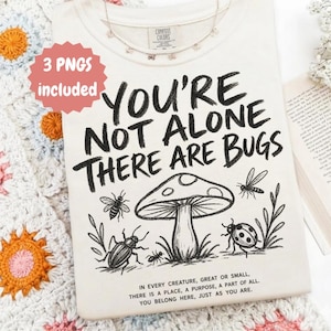 May include: A cream-colored t-shirt featuring black line art of a mushroom, a beetle, a ladybug, and a bee. The text reads: YOU'RE NOT ALONE THERE ARE BUGS and IN EVERY CREATURE, GREAT OR SMALL, THERE IS A PLACE, A PURPOSE, A PART OF ALL.
