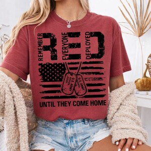K&ouml;nnte beinhalten: Ein weinrotes T-Shirt mit den W&ouml;rtern "RED" und "DEPLOYED" in gro&szlig;en, fetten, schwarzen Buchstaben. Das Shirt zeigt ein amerikanisches Flaggen-Design und zwei Erkennungsmarken. Der Text "REMEMBER EVERYONE DEPLOYED UNTIL THEY COME HOME" ist ebenfalls aufgedruckt.