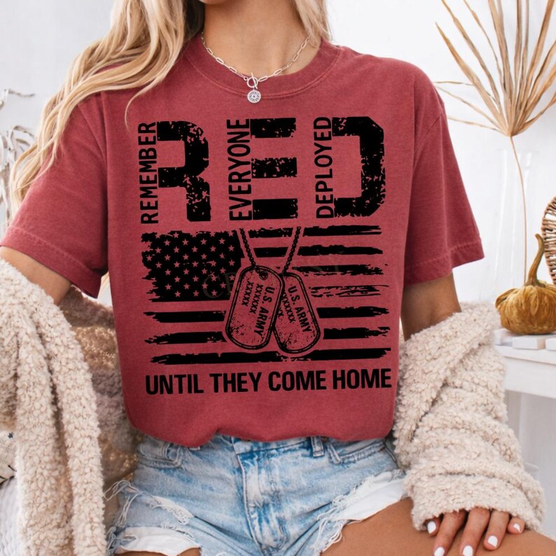 K&ouml;nnte beinhalten: Ein weinrotes T-Shirt mit den W&ouml;rtern "RED" und "DEPLOYED" in gro&szlig;en, fetten, schwarzen Buchstaben. Das Shirt zeigt ein amerikanisches Flaggen-Design und zwei Erkennungsmarken. Der Text "REMEMBER EVERYONE DEPLOYED UNTIL THEY COME HOME" ist ebenfalls aufgedruckt.