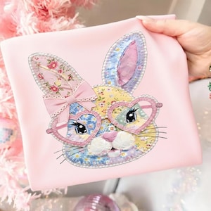 Könnte beinhalten: Ein rosa Stoffartikel mit einem Patchwork-Hasengesicht-Design. Der Hase hat herzförmige Brillen und eine Schleife, mit floralen und gemusterten Details an Ohren, Gesicht und Brille. Der Hintergrund ist ein zartes Rosa.