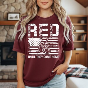 K&ouml;nnte beinhalten: Ein weinrotes T-Shirt mit einem verblassten Design der amerikanischen Flagge. Das Shirt zeigt die Worte "REMEMBER EVERYONE DEPLOYED" in wei&szlig;en Blockbuchstaben, zusammen mit zwei Erkennungsmarken und dem Satz "UNTIL THEY COME HOME."