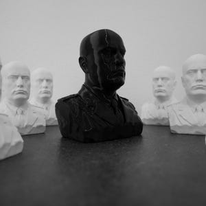 Puede incluir: Un busto central negro brillante de un hombre con uniforme militar rodeado de varios bustos blancos mate de la misma figura. Cada busto representa a un hombre calvo con expresión severa, vistiendo camisa con cuello, corbata y chaqueta con charreteras.