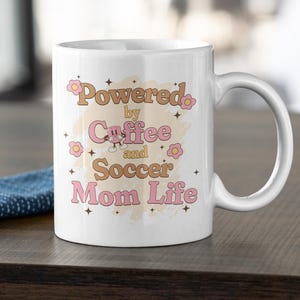 May include: White ceramic mug with the text "Powered by Coffee and Soccer Mom Life" in pink and brown lettering. The mug has a curved handle and is decorated with small flower graphics.