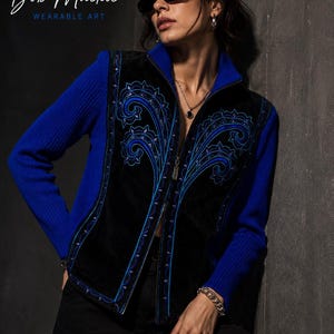 May include: A blue zip-up sweater with a black velvet vest, adorned with blue embroidered floral patterns. The vest has a zip closure. The text "Bob Mackie WEARABLE ART" is visible. The overall design suggests a focus on wearable art.