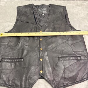 May include: A black leather waistcoat featuring a patchwork design, a V-neckline, and five brass-coloured snap buttons. A yellow ruler placed across the chest shows a width of approximately 61 cm. The interior label reads Genuine Leather.