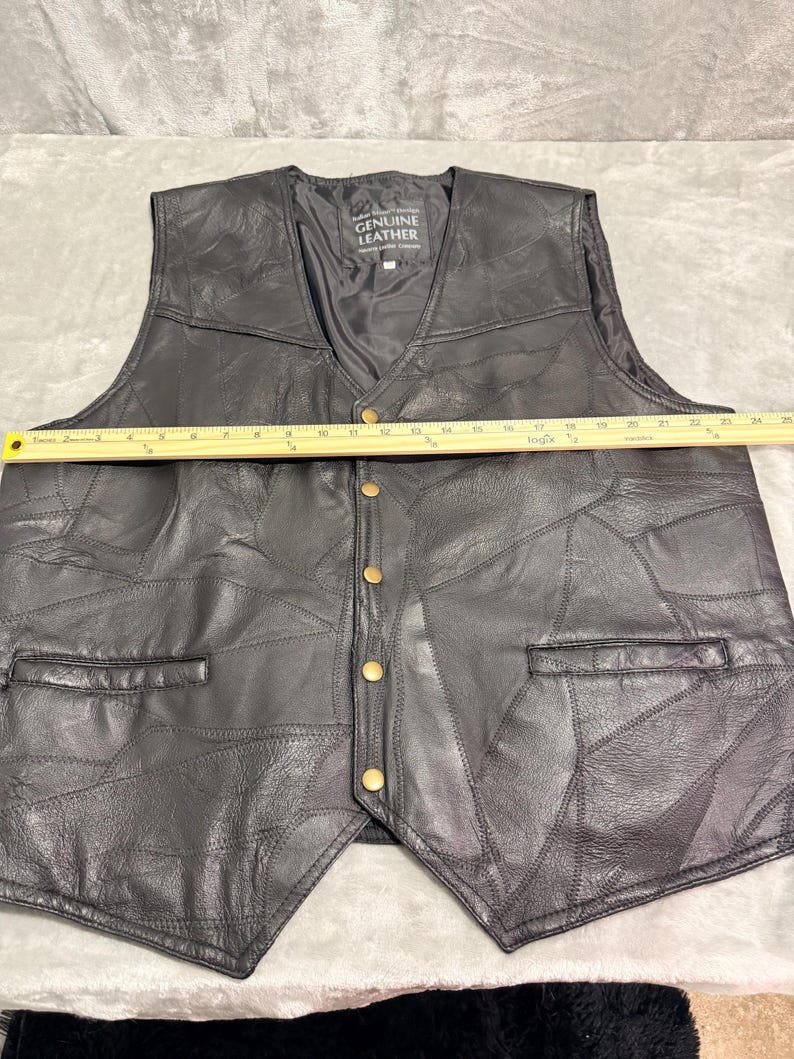 May include: A black leather waistcoat featuring a patchwork design, a V-neckline, and five brass-coloured snap buttons. A yellow ruler placed across the chest shows a width of approximately 61 cm. The interior label reads Genuine Leather.