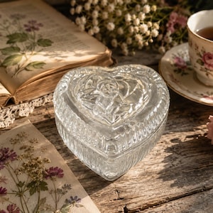 May include: A clear glass trinket box shaped like a heart with a removable lid. The lid features a moulded rose design, and the sides of the box have vertical ribbed detailing. The glass is transparent and reflects light across its textured surface.
