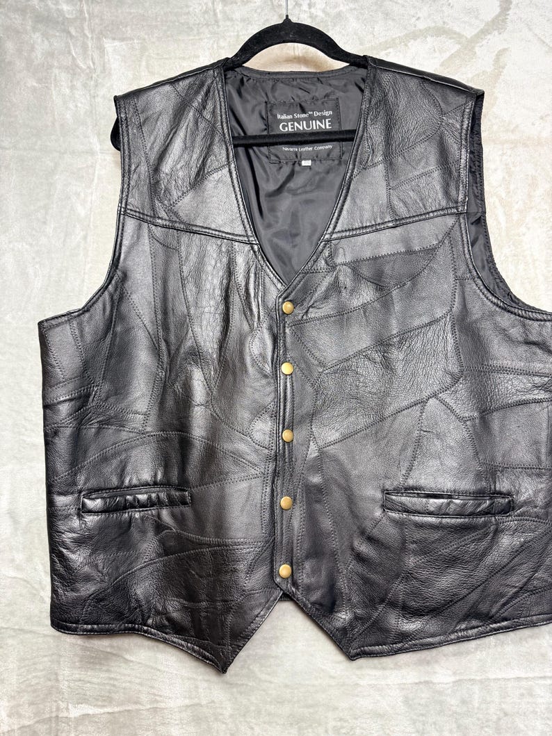 May include: A black leather waistcoat featuring a patchwork design of various leather pieces. The waistcoat has a V-neckline, five gold-toned metal snap buttons down the front, and two horizontal slit pockets. A label inside reads Italian Stone Design GENUINE.