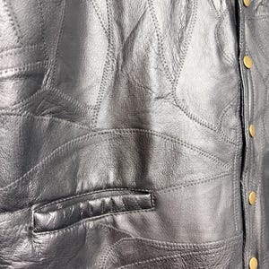 May include: A black leather waistcoat featuring a patchwork design with visible stitched seams throughout the garment. The waistcoat includes a horizontal welt pocket on the front and a vertical placket secured with five round, gold-toned metal snap buttons.