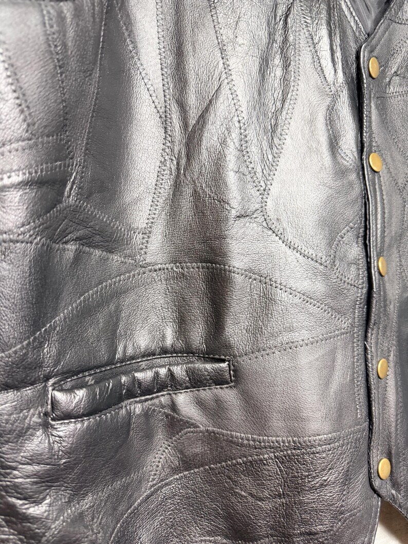 May include: A black leather waistcoat featuring a patchwork design with visible stitched seams throughout the garment. The waistcoat includes a horizontal welt pocket on the front and a vertical placket secured with five round, gold-toned metal snap buttons.