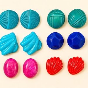 May include: A collection of twelve buttons arranged in pairs, featuring various shapes and colours including teal fan-shaped, green textured round, light blue shell-shaped, dark blue faceted round, pink marbled oval, and red shell-shaped buttons.