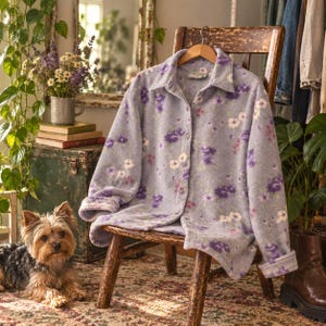 May include: A long-sleeved button-down shirt in a light lavender fleece fabric featuring an all-over floral pattern in shades of purple, white, and soft pink. The shirt has a classic collar, a front button placket, and cuffed sleeves.