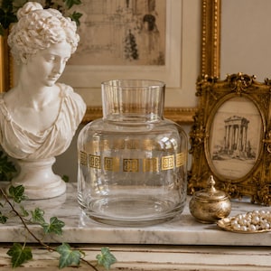 May include: A clear glass vase featuring a gold-coloured Greek key pattern around its midsection. The vase has a wide, rounded body with a shorter, straight neck and an open top, designed for holding floral arrangements or decorative displays.