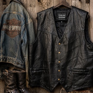 May include: A black leather waistcoat with a patchwork design, featuring a V-neckline, five brass-coloured buttons, and two front pockets. The interior label reads Genuine Leather. Beside it hangs a blue denim jacket with a Harley-Davidson logo patch.